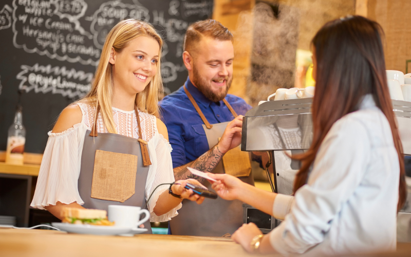 A barista hands back a payment card to a customer while another barista operates an espresso machine in a café.