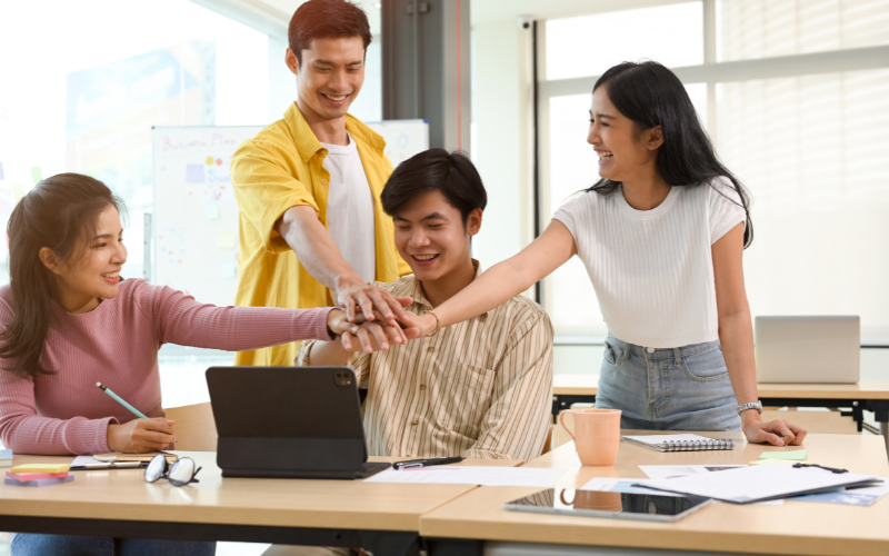 Four people standing and sitting at a table, placing their hands together in a group gesture, smiling, with a whiteboard in the background.