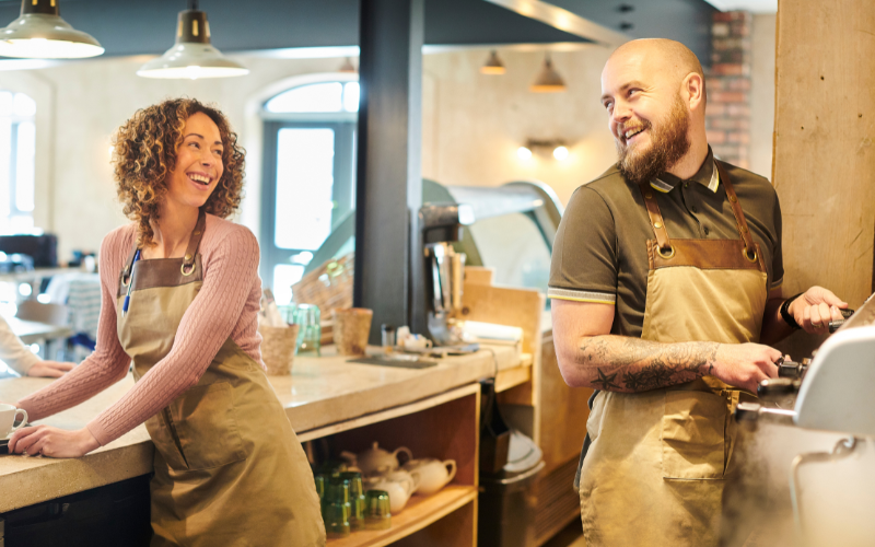 Two smiling baristas wearing aprons interact behind the counter in a cafe, with coffee equipment and supplies in the background.