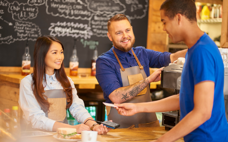 Two baristas, one male and one female, serve a customer at a café counter. Chalkboard menus are visible in the background.