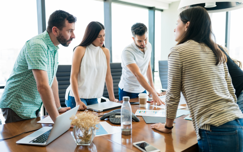 Four people are engaged in a discussion around a conference table with documents, laptops, and coffee cups present.