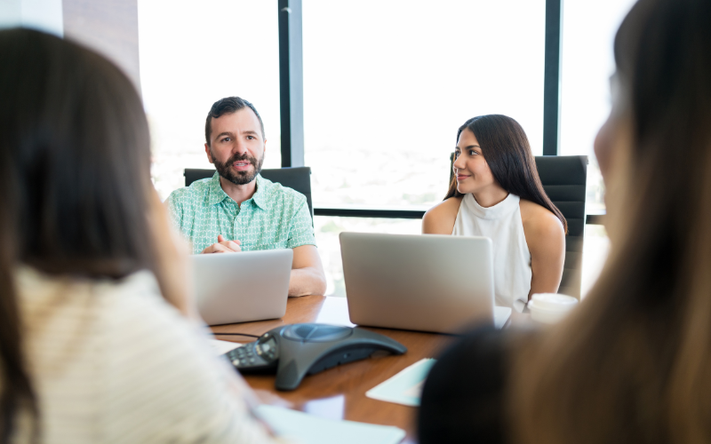 A group of people are sitting around a conference table with laptops, engaging in a discussion in a well-lit office.