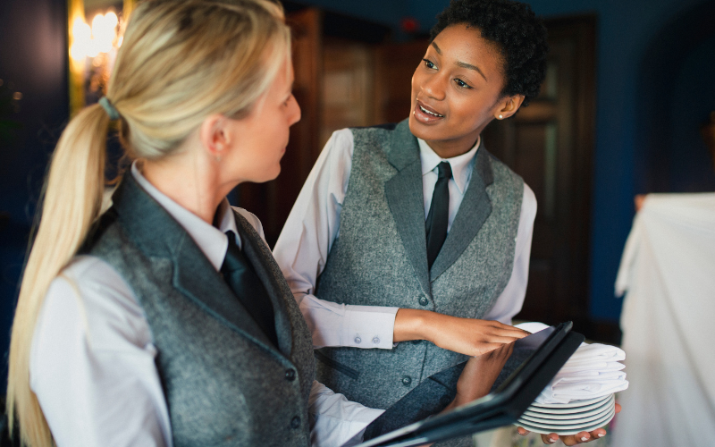 Two women in uniform vests are discussing while holding plates and a tablet. They are to be working in a hospitality setting.