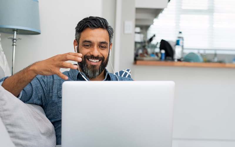 Man with earbuds smiling during a Telehealth video call on a laptop while sitting on a sofa in a well-lit room.