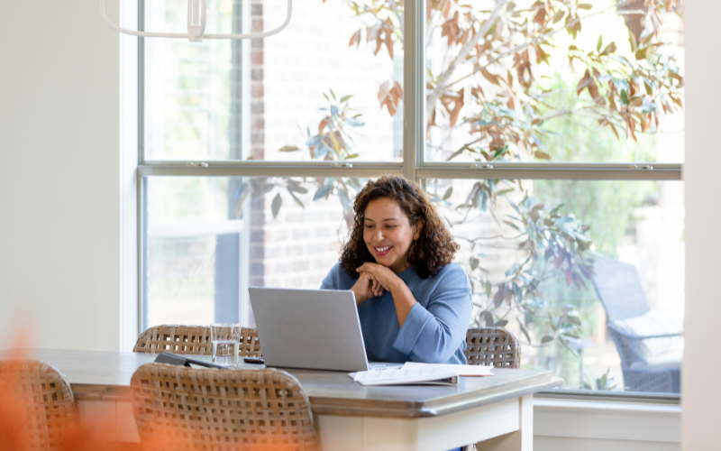 A female therapist in a blue sweater sits at a table, using a laptop for Telehealth, with a large window and trees in the background.