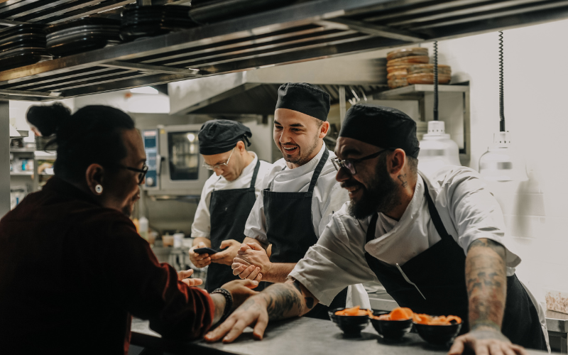 Four chefs in a restaurant kitchen, two of them smiling and engaging with a manager at the counter.