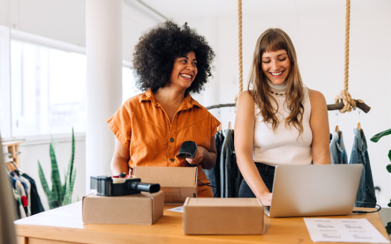 Two women productively and happily at a workspace with a laptop, boxes, and a barcode scanner on a wooden table.