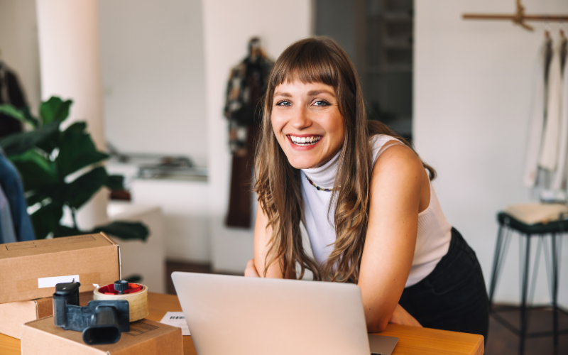 An employee of a small business in a white sleeveless top smiles while leaning on a table with a laptop and boxes in a room with clothes in the background.