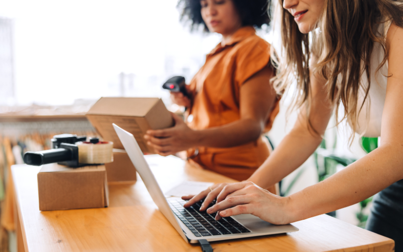 Two women working at a small business; one uses a barcode scanner on packages, the other types on a laptop on a wooden desk.