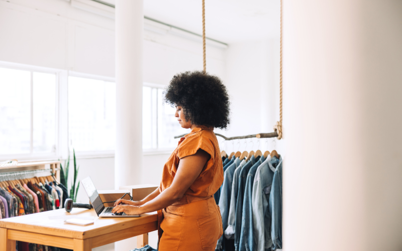 Person in an orange outfit working on a laptop at a desk in a clothing store with racks of clothes in the background.