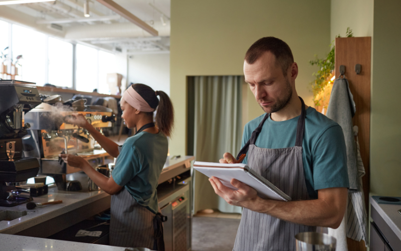 Two café workers in aprons: one taking notes, the other operating an espresso machine. Bright, modern café setting.