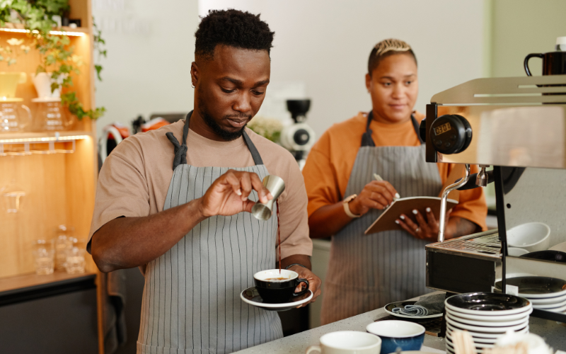 Two baristas preparing a coffee drink at a cafe, with one pouring milk and the other taking notes beside an espresso machine.