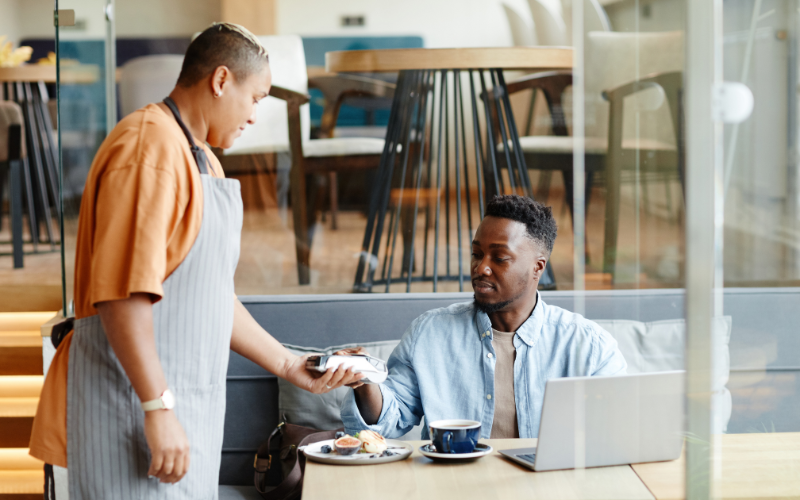 A person hands a bill to a seated man at a local café table with a laptop, coffee, and dessert in a modern setting.