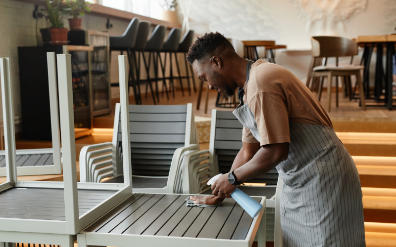 Person cleaning a table in a restaurant, wearing a striped apron and using a spray bottle and cloth.