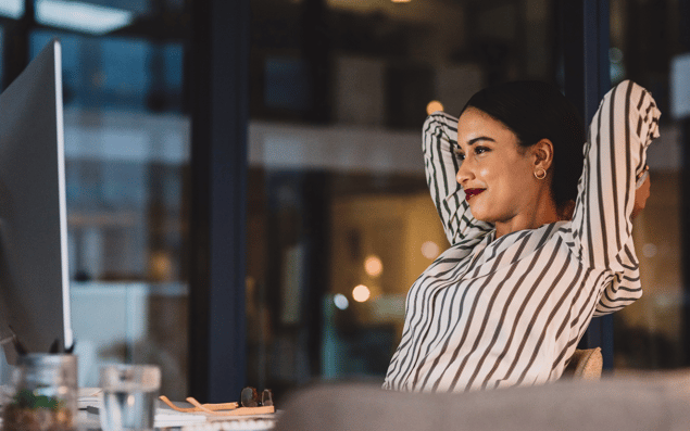 A woman in a striped shirt relaxes with hands behind her head while looking at a computer screen in a dimly lit office.