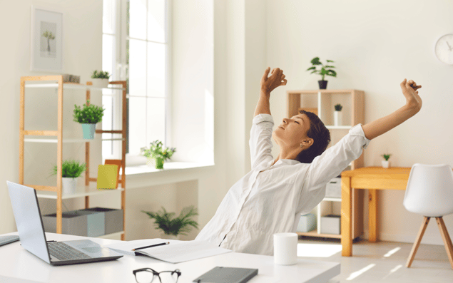 Person stretching with arms raised in a home office, sitting at a desk with a laptop, glasses, and potted plants in the background.