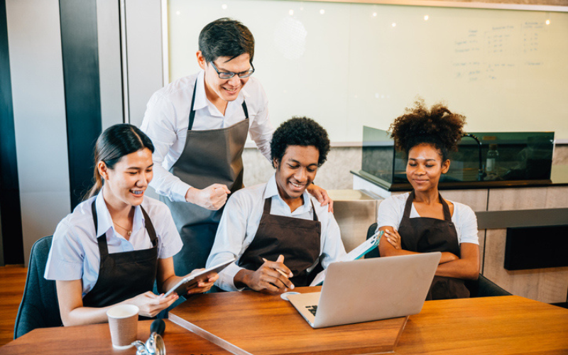 Four team members in uniforms gather around a laptop in a break room in a happy work place culture.