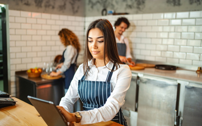 Woman in striped apron working, using tablet in a restaurant, while two people work in the background with food.