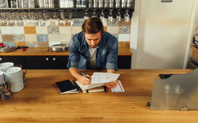 Business owner in a denim shirt works on paperwork at a wooden counter in a cafe setting, with shelves and cups in the background.