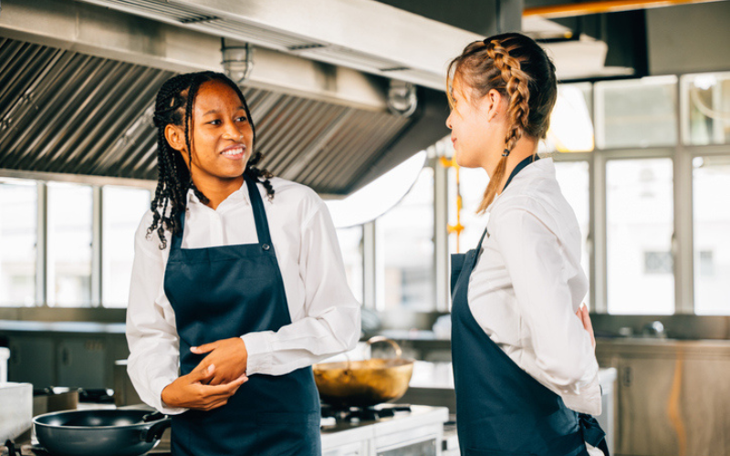 Two people in aprons converse in a professional kitchen, surrounded by cooking equipment and stovetops.