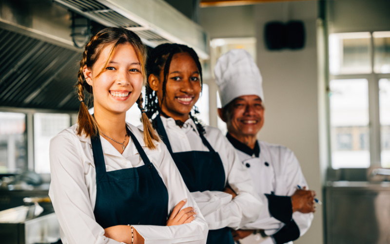 Three employees in uniform and aprons stand smiling in a kitchen setting.