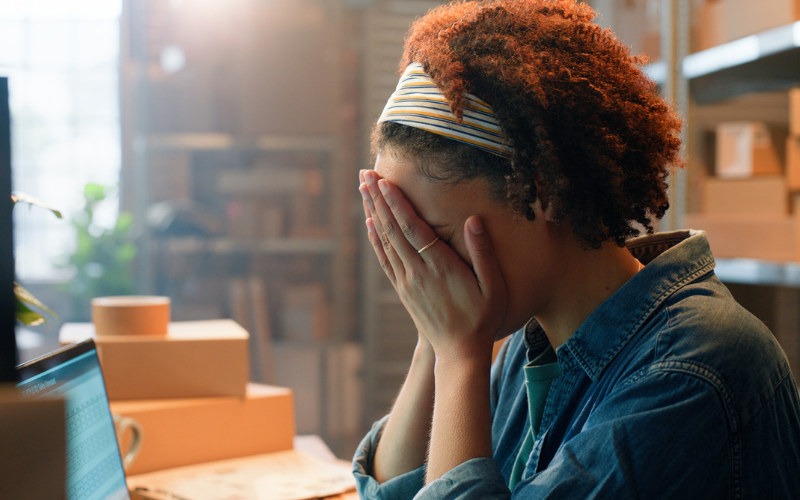 A person sits at a desk with hands covering their face, surrounded by boxes and a laptop in a workspace.