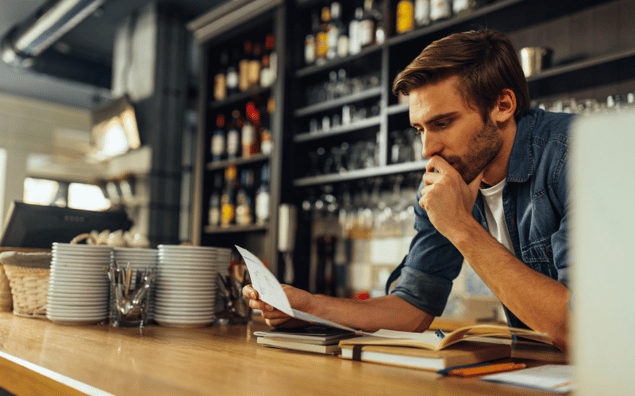 business owner in a cafe reviews a document while resting his chin on his hand, surrounded by books and dishes on the counter.