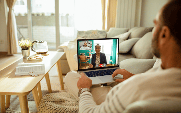 Person sitting on a couch having a telehealth therapy dog on a laptop with two people visible on the screen.