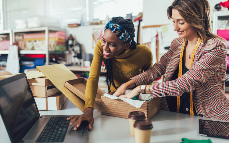 Two women packing a cardboard box working together, emphasizing teamwork and employee well-being in a retail environment.