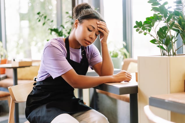 An employee wearing an apron sitting at a table in a café, looking tired and burnt-out with one hand on their forehead.