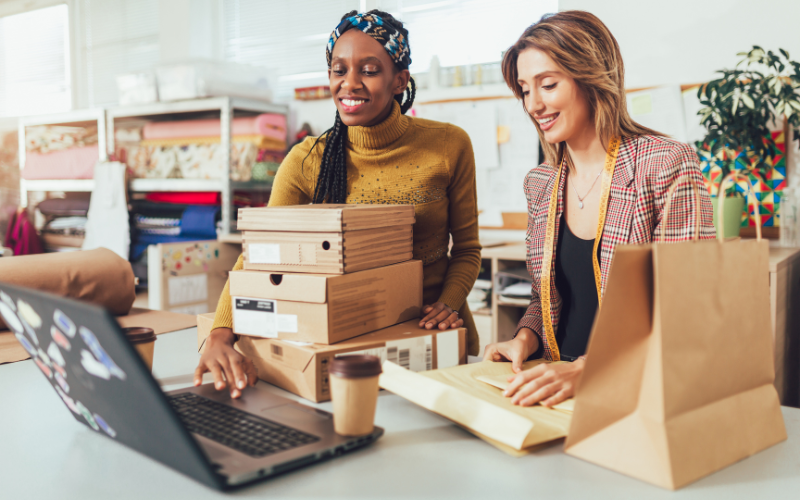 Two women working at a table with cardboard boxes and a laptop, preparing packages for shipping in a retail store.