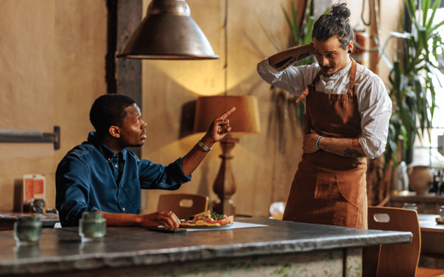 A man sitting at a table points at a standing server in an apron inside a rustic room with plants and a lamp.