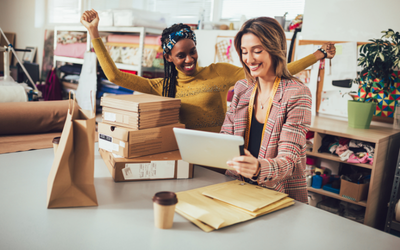 Two women celebrate at a retail store with stacks of packages on the table as one holds a tablet and the other raises her arms in joy.