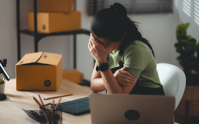 small business owner sitting at a desk with head in hand, surrounded by boxes and office supplies, appearing stressed.