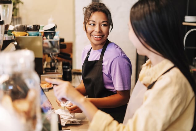 Two women wearing aprons are smiling and conversing behind the counter at a coffee shop. One is holding a receipt.