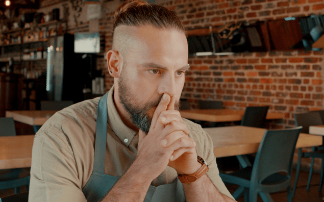 An employee with a beard sits at a table in a cafe, looking thoughtful, with his hands folded near his face.