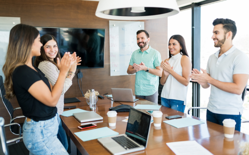 Five people stand around a conference table in a modern office, clapping and smiling during a meeting. Laptops and coffee cups are on the table.