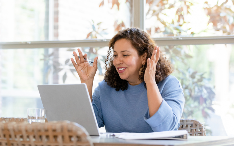 A female therapist in a blue sweater sits at a table, using a laptop for Telehealth, with a large window and trees in the background.