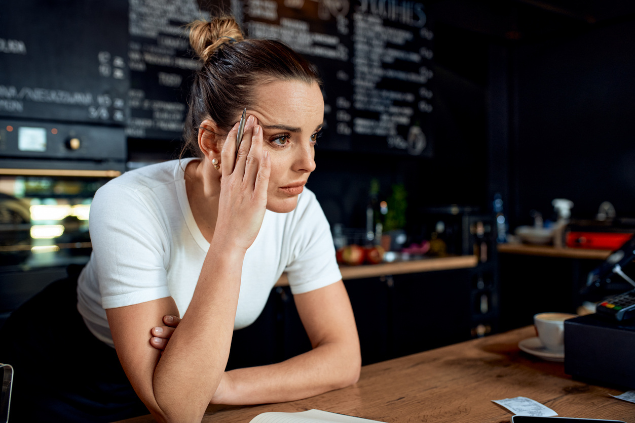 A woman in a white shirt looks stressed while leaning on a counter in a café with a menu board in the background.